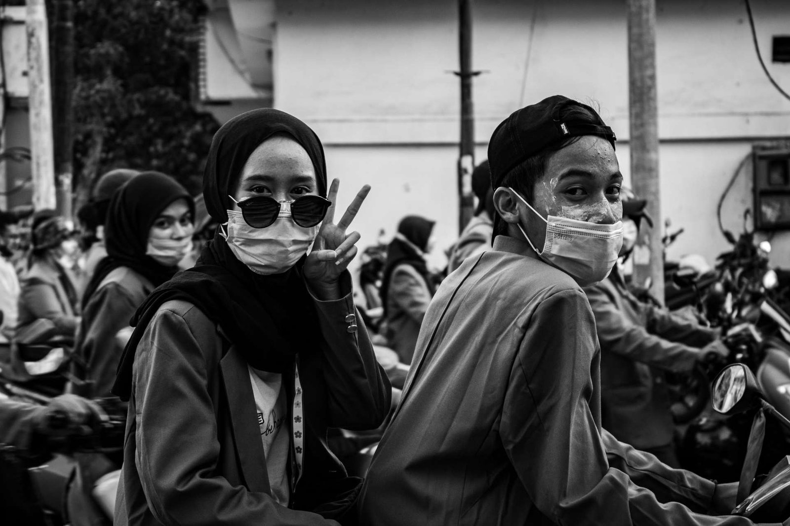 Two young people in masks on a motorbike in a crowd, one holding up a peace sign.