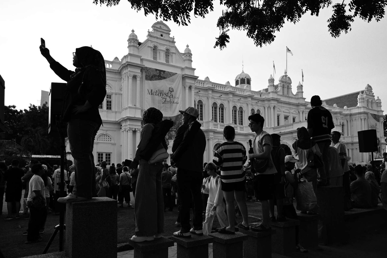 Silhouetted crowd standing on concrete blocks, one person holding up a phone, in front of a white colonial building.